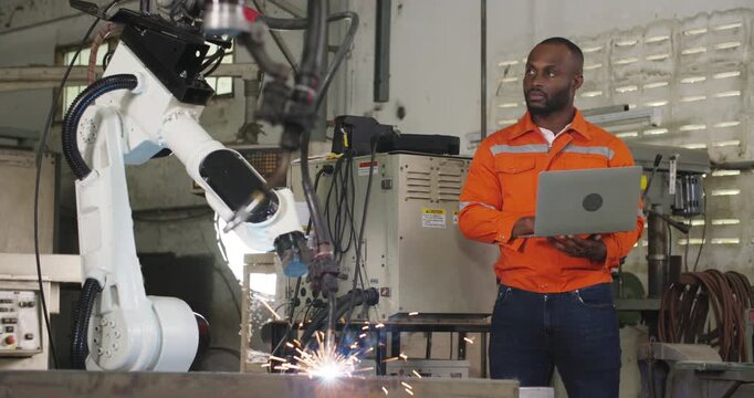 Professional industrial engineer African american man using computer laptop and to control and programing automation robot arms machine in manufacturing industry. Technology and innovation concept.