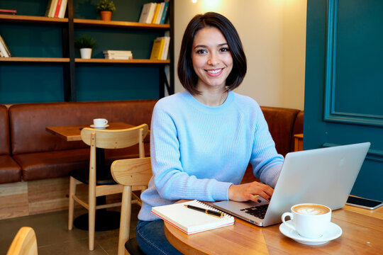 Portrait of young Asian woman working on laptop in cafe, smiling at camera while typing and planning tasks. Suitable for remote work, freelance business, digital productivity marketing