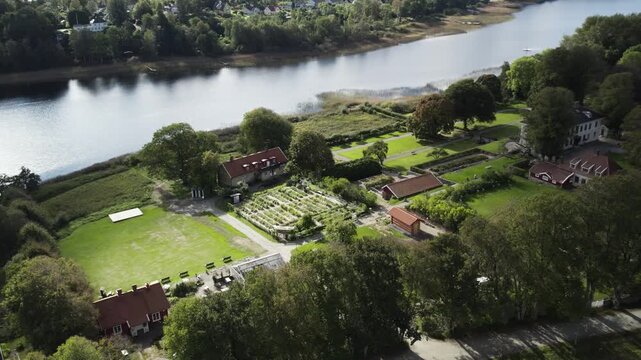 Aerial drone view of Rada Sateri manor gardens beside Radasjon Lake in Harryda. Formal garden plots, open lawns and surrounding buildings overlook calm lakeshore and wooded landscape