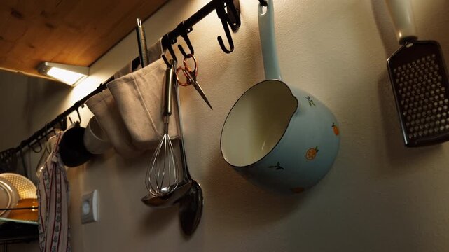 Saucepan, whisk, ladle, grater and other utensils hang from hooks on a kitchen wall, showing organized cookware and everyday home meal preparation.
