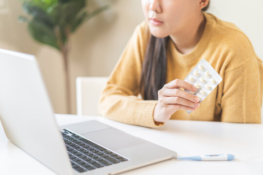 Asian young woman holding a blister pack of pills during an online medical consultation with laptop and thermometer on table, home healthcare and telemedicine concept.