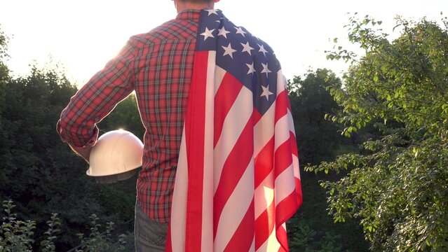 Construction worker holds a white hard hat with an American flag draped over his shoulder. Symbolizes American workers or Labor Day. Concept of pride in construction profession and American dream