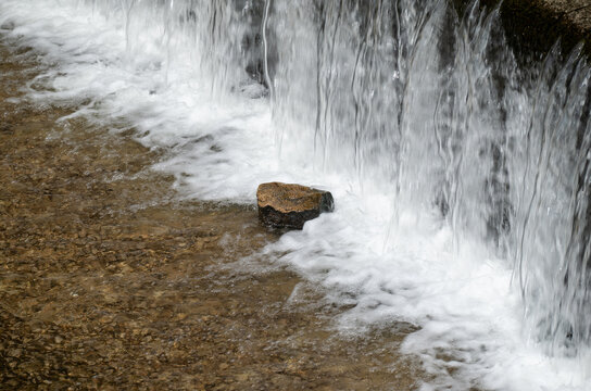 Close-up of crystal clear water cascading over a small dam with a single rock in the bubbling stream