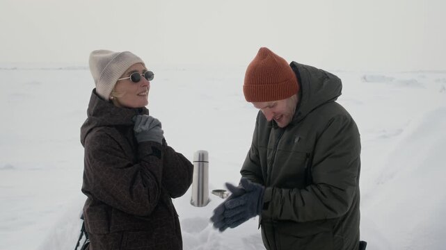 Caucasian adult couple standing in snowy landscape, warming gloved hands and talking near thermos suitable for winter tourism and outdoor apparel marketing