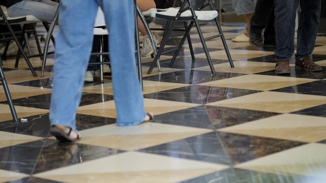 Checkered marble floor with black and beige squares. People walk past folding chairs. Legs and feet move quickly, no visible emotions. Busy indoor scene, neutral atmosphere