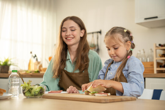 Little girl holding an open-face sandwich with ham on her palm while chatting with her mother. Happy family cooking. Cute daughter showing her homemade sandwich to her smiling mom in the kitchen.