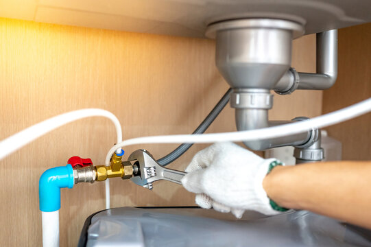Plumber hand with glove fixing pipe connection with adjustable wrench and checking grease trap box under kitchen sink. Handyman repairing plumbing system in wooden cabinet.