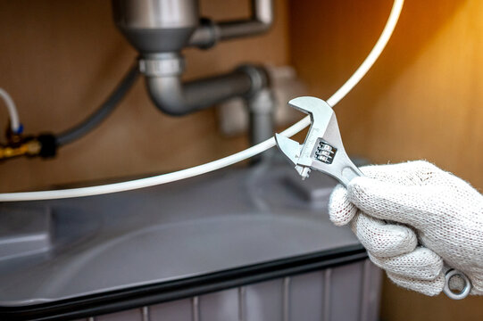Plumber hand with glove fixing pipe connection with adjustable wrench and checking grease trap box under kitchen sink. Handyman repairing plumbing system in wooden cabinet.