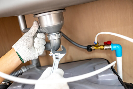 Plumber hand with glove fixing pipe connection with adjustable wrench and checking grease trap box under kitchen sink. Handyman repairing plumbing system in wooden cabinet.