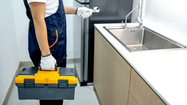 Male plumber with gloves holding yellow toolbox and wrench in the kitchen. Handyman in uniform prepares to fix plumbing system under stainless steel sink in wooden cabinet. Home maintenance service