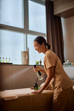 Smiling Korean wellness therapist spraying massage table with aromatic scent before treatment.