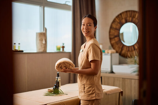 Happy  Korean massage therapist arranging towels in spa room and looking at camera.