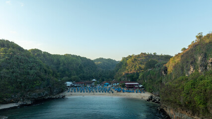 Fototapeta premium Aerial view of a secluded turquoise cove surrounded by rugged forested cliffs along the tropical Ngerenean coastline in Indonesia.