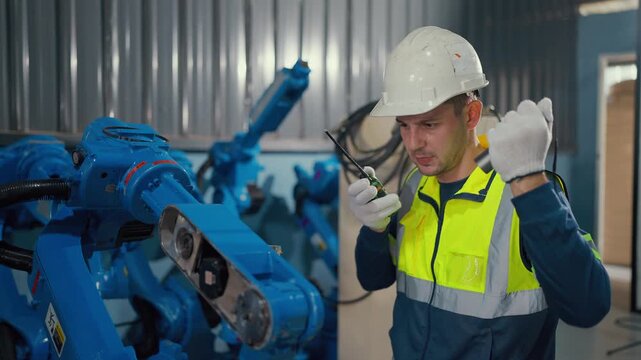 Technician checks robotic arm in factory setting. Worker wears safety gear and white helmet. He focuses intently on diagnostic tools