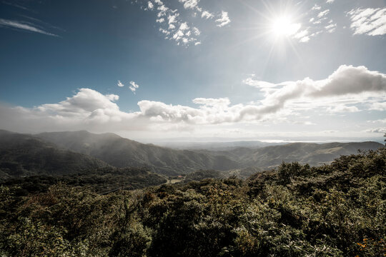 Panoramic sunlit valley with rolling green hills and ridges, mist-filled lowlands, distant mountains and dramatic clouds over landscape