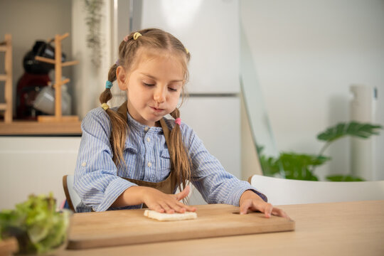 Cute little girl pressing a bread slice on wooden cutting board while making sandwich in kitchen. Adorable caucasian child enjoying preparing healthy breakfast snack at home kitchen table.