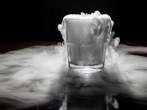 A glass filled with thick white vapor overflows and swirls across a dark, reflective surface, creating a dramatic and mystical atmosphere.