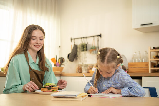 Caucasian little girl doing homework at kitchen table while mother bringing snack. Cute focused daughter writing in notebook with mother holding a sandwich plate in background.