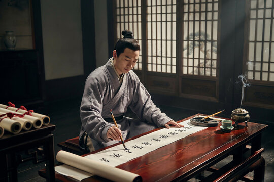 A man in traditional chinese robes sits on the floor at a low wooden table, practicing calligraphy on a long scroll with a brush and ink