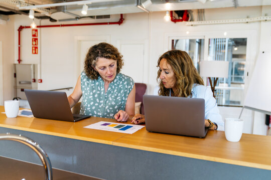 Two businesswomen discussing data charts, using laptops, and working together in a modern, shared office environment