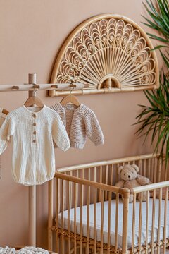 Softly blurred view of cream muslin romper and knit cardigan on wooden peg rail near natural rattan crib with plush teddy bear inside