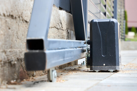 Automatic sliding gate motor mechanism installed on concrete base next to gray metal fence provides secure entrance control, modern house protection, automated private property access.