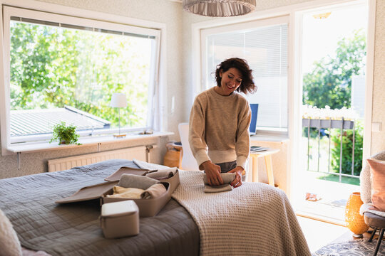 Smiling mature woman rolling clothes on bed in bedroom