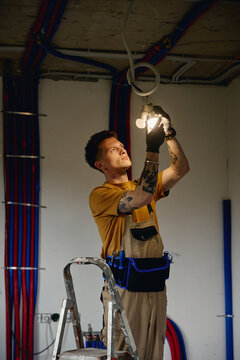 Vertical image of Caucasian young adult male electrician installing light bulb on ceiling during house renovation, suitable for home improvement and construction services marketing