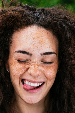 Cheerful young curly haired woman with eyes closed sticking out tongue