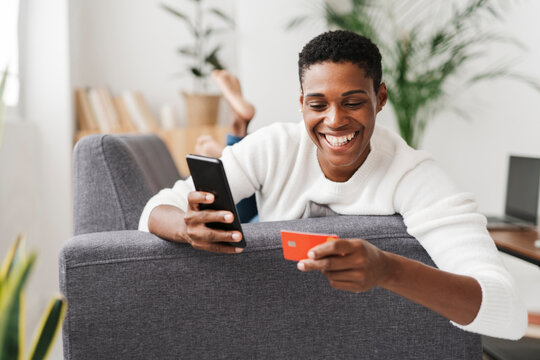 Smiling woman lying on couch using mobile phone and credit card