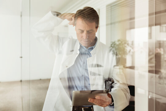 Mature man in lab coat using digital tablet behind glass pane