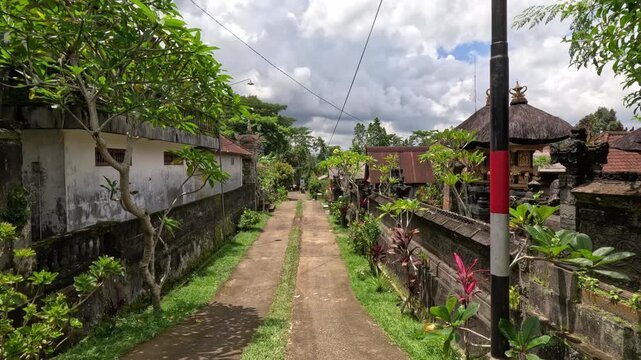 Wide shot of a residential street on Bali island during a sunny day, showing houses with dedicated Balinese shrines, lush greenery, and a clear blue sky.