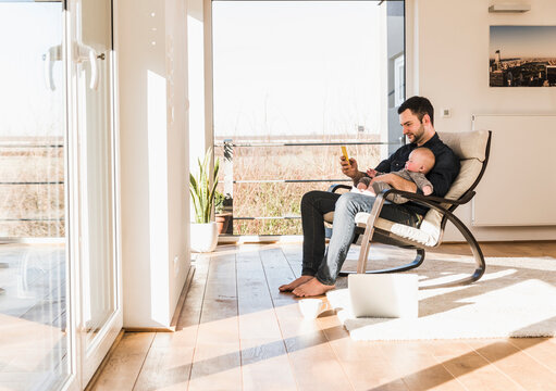 Father sitting in arm chair with baby son on lap, using mobile devices