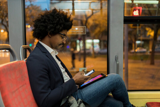 Spain, Barcelona, businessman in a tram at night using cell phone and tablet