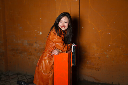 Smiling model in red raincoat leaning on box in city evening