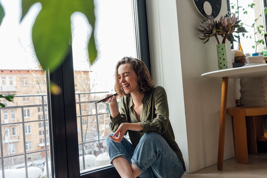 Cheerful woman with eyes closed sending voicemail through smart phone by glass window at home