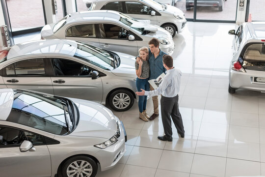Car dealer showing new car to young couple in showroom