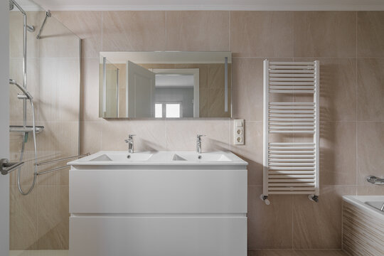 Modern neutral bathroom interior with double sink, large mirror, chrome faucets, towel radiator, beige tiles, and sleek minimalist finishes creating bright spacious comfort