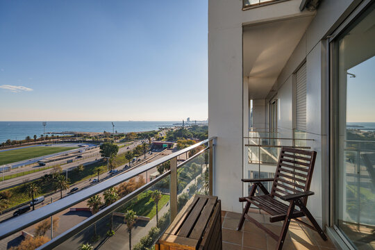 Modern balcony with wooden chair overlooking coastal ocean and cityscape beneath clear blue sky, featuring glass railing, sunlit terrace, urban horizon and relaxed seaside living