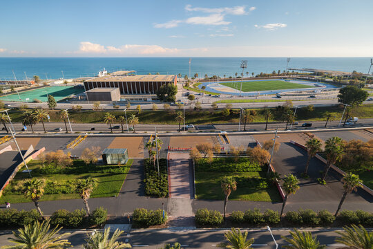 Aerial view coastal park with palm trees, sports fields, landscaped gardens, waterfront buildings, marina, blue ocean horizon, clear sky, and modern city shoreline architecture