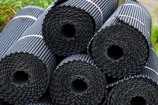 Stacked rolls of black dimpled HDPE drainage membrane stored at a construction site. A close-up cross-section view highlights the spiral pattern and textured dimpled surface used for foundation waterp