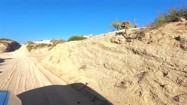 Window mounted shot from a car driving along the East Cape road in Baja California Sur, dropping down into a small arroyo where a brief view of the ocean and a house on the far side appear before disa