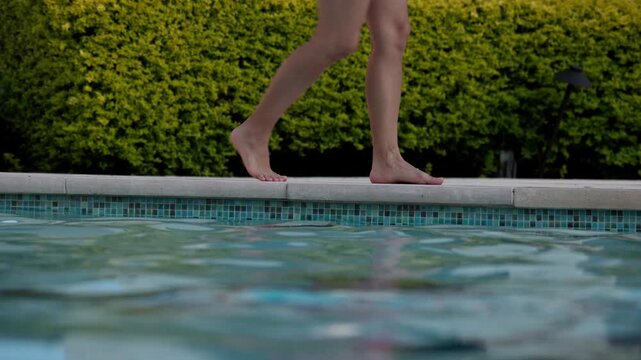Woman walking in slow motion past the edge of a swimming pool as it ripples against a green bush background