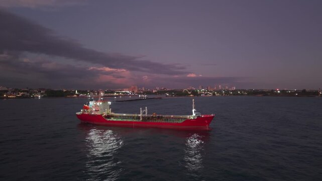 An aerial drone shot of a red cargo ship sailing through coastal waters at dusk, with the illuminated cityscape glowing softly along the distant horizon