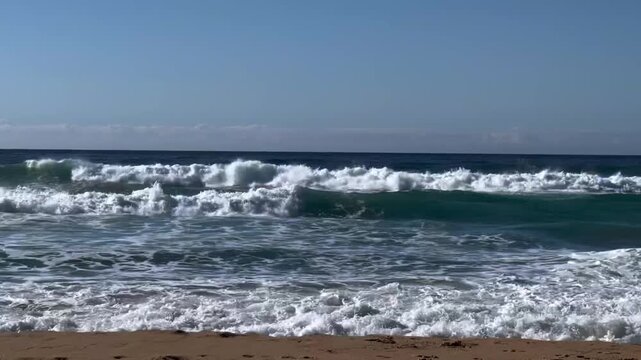 Ocean waves rolling and crashing on sandy beach shore under clear blue sky during bright daylight