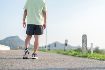 Fototapeta premium Young adult Asian man jogging and walking in the park at morning. male athlete in sport shoes running on the road with visible leg muscles. Healthy lifestyle, wellness and workout concept