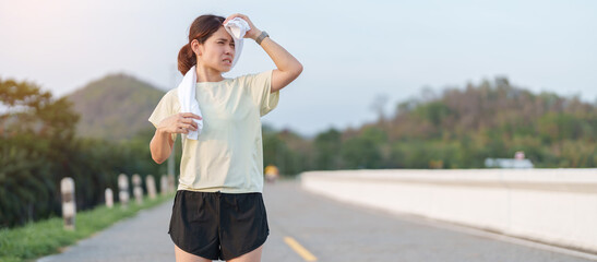 Fototapeta premium Exhausted Asian woman wiping sweat with a towel after jogging in the park. Tired female runner suffering from summer heat and fatigue after fitness workout. Outdoor exercise and heatstroke concept