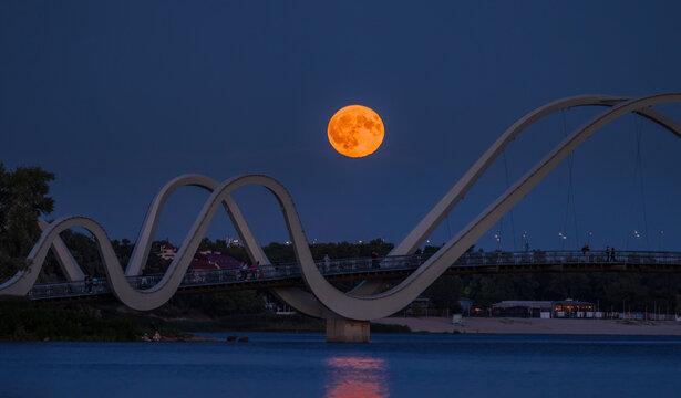 Full Buck Moon rising behind the Wave pedestrian bridge over the Dnipro River in Obolon, Kyiv, Ukraine. Glowing orange moon framed by modern architecture at blue hour.