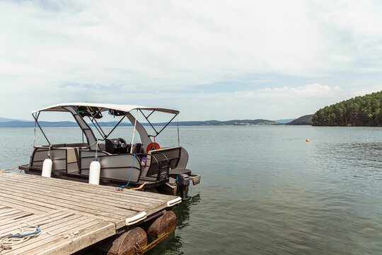 Modern pontoon boat with canopy docked at a weathered wooden pier on a calm lake, with distant forested islands and a cloudy sky, perfect for leisure boating and water recreation.
