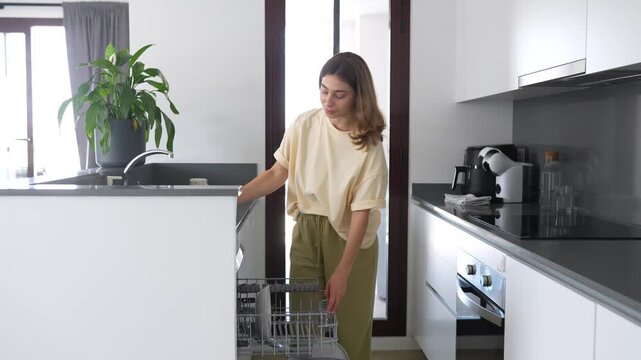 Woman loads dirty dishes into dishwasher in a modern kitchen. Housewife cleans a bright kitchen, disassembling the dishwasher. Concept of cleanliness, cleaning, and technology.